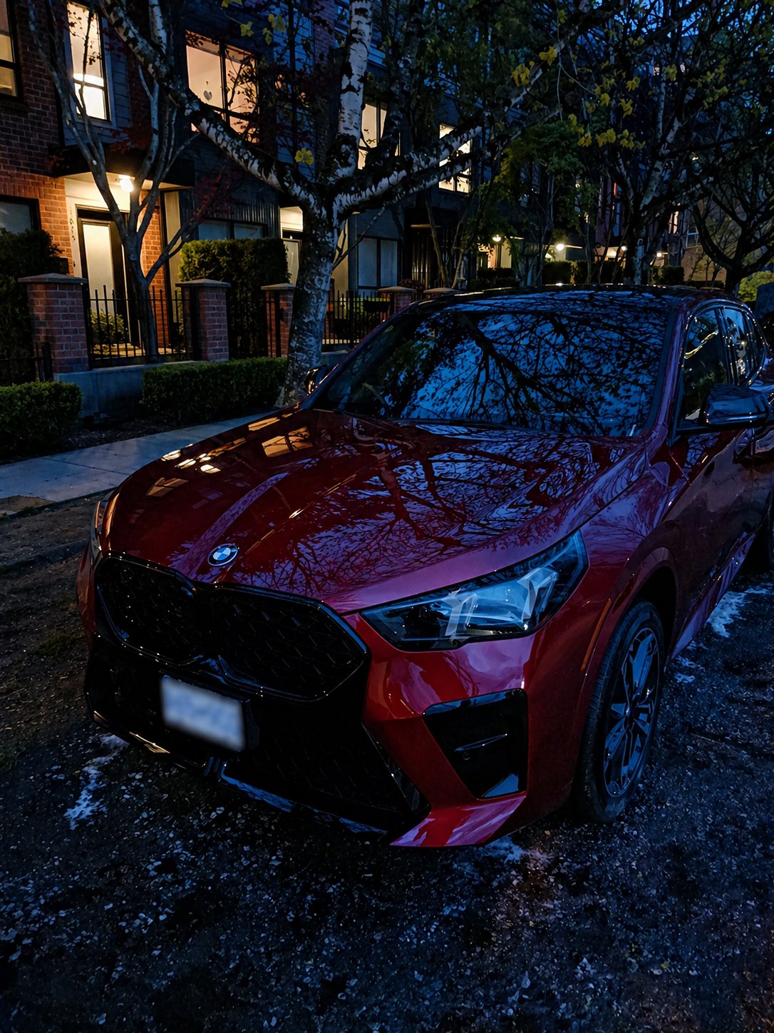 Red car parked on suburban street at dusk with illuminated house and trees in background
